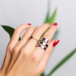 Close-up of a hand wearing two rings: a slim gold ring with a small brown gemstone and a large silver stacked ring, red nails, and green leaves in the blurred background.