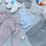 Flat lay of baby essentials on a blue background: striped onesie, gray shorts, blue socks, bottle, pacifier, rattle, and teether.