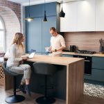 Woman sits at a wooden kitchen island with a laptop, while a man pours coffee nearby in a modern kitchen.