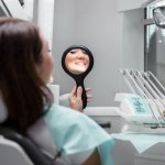 Patient in a dental chair smiles while holding a handheld mirror to view her teeth in the mirror frame.