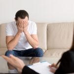 Man on a beige couch covers his face with his hands during a therapy session.