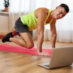 Man in a neon yellow tank top on a pink yoga mat performs a hand‑and‑knee exercise in a bright living room, looking at a laptop nearby.