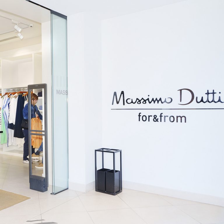 772741 - Outlet Social - Revista DeBelleza.Es Massimo Dutti store entrance with white walls, glass door, and a shopper browsing clothing inside.