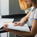 Person wearing a light uniform studying at a desk, turning pages in a book with a notebook and pencil nearby.