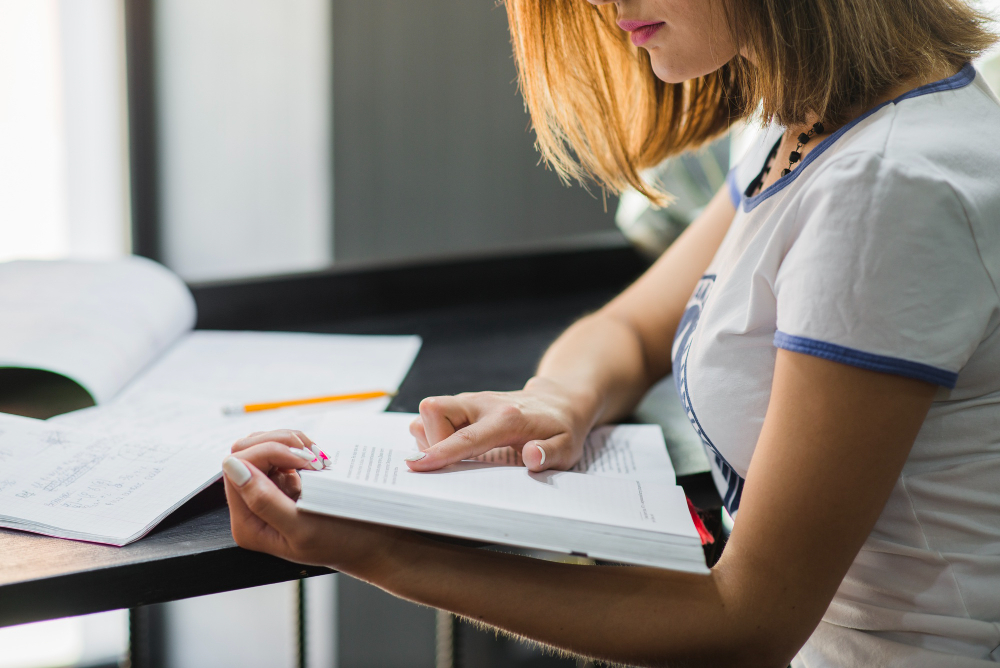 772920 - SacaOpos - Revista DeBelleza.Es Person wearing a light uniform studying at a desk, turning pages in a book with a notebook and pencil nearby.