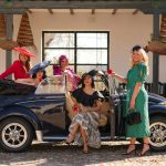 Group of stylish women in vintage dresses and hats posing with a navy convertible in a sunny courtyard.