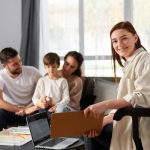 Therapist with clipboard smiles at camera as a family sits on a sofa in a bright living room.