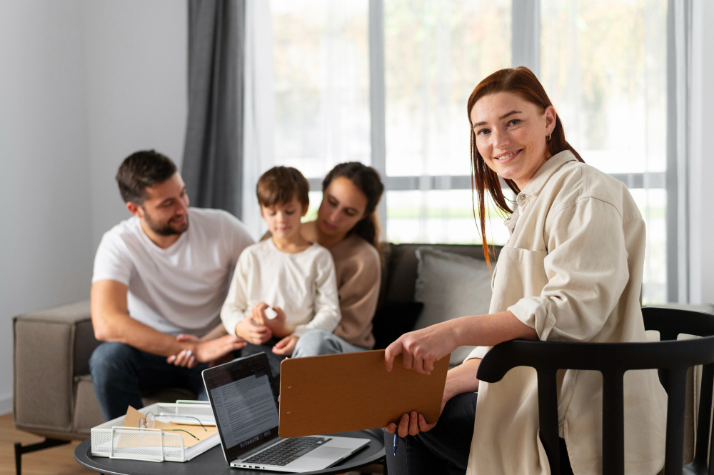 Therapist with clipboard smiles at camera as a family sits on a sofa in a bright living room.