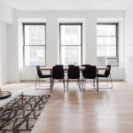 Bright modern dining room with a long wooden table and eight black chairs by large windows; a gray sofa on the left.
