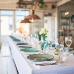 Long banquet table set for a meal with green plates, glassware, and white linens in a bright, airy room.