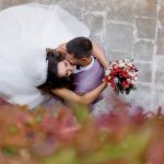 Top-down view of a bride and groom embracing, the bride in a white wedding gown with veil and a pink-red bouquet in focus.
