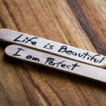 Two wooden sticks with handwritten inspirational phrases: 'Life is Beautiful' and 'I am Perfect' on a wooden surface.