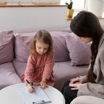 Young girl drawing with a marker on a clipboard while an adult woman sits beside her on a lilac sofa in a bright living room.