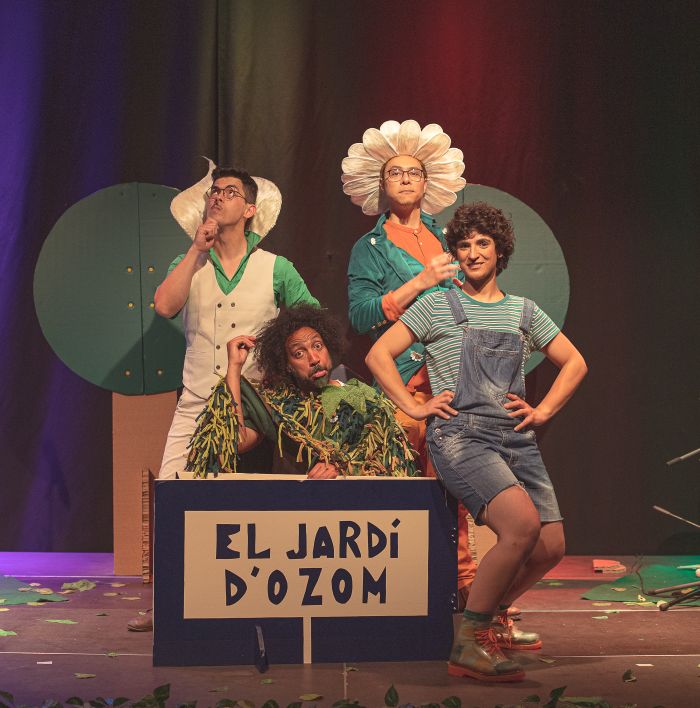 Theater stage with four performers in colorful garden costumes posing around a blue prop box labeled 'EL JARDI D'OZOM'. One wears a sunflower hat.
