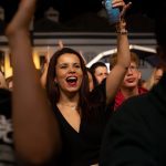 Joyful woman in a black outfit raises a blue cup while cheering in a crowded nighttime event.