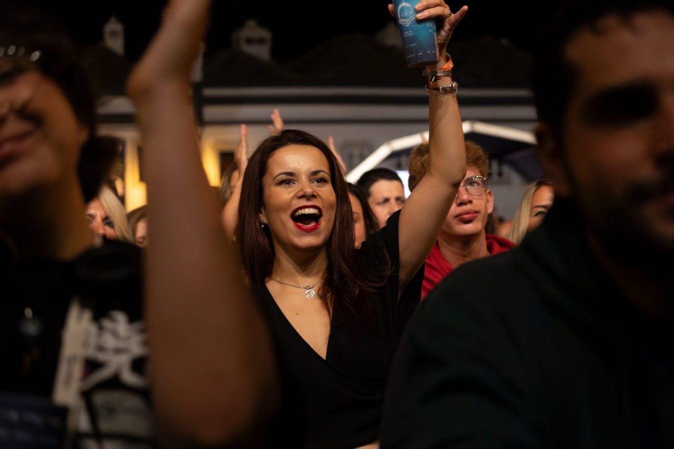 775623 Eventos 981 - Revista DeBelleza.Es Joyful woman in a black outfit raises a blue cup while cheering in a crowded nighttime event.