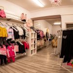 Interior of a children's clothing store with racks of colorful outfits along the left wall, and a rack of black pants on the right; warm lighting and a decorative chandelier overhead.