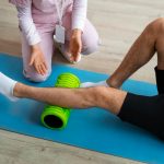 Therapist assisting a person performing leg foam rolling on a blue exercise mat with a bright green roller under the shin area.