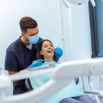 Dentist in a mask and blue gloves performing a dental checkup on a patient seated in a dental chair.