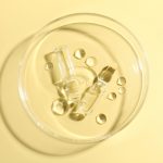 Two clear glass ampoules filled with liquid sit in a shallow glass dish with scattered droplets on a pale yellow background (cosmetic/serum theme).