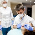 Dentist in mask and gloves performs a dental procedure on a patient, with an assistant in the background in a bright clinic.
