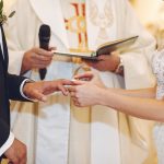 Bride and groom exchange rings during a wedding ceremony, officiant with a book in the background