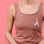 Person wearing a dusty-rose tank top with a pink breast cancer awareness ribbon pinned to the chest against a pink background